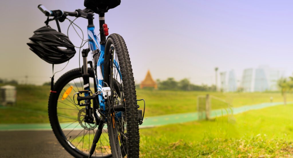 Endlich Frühling! Ein Fahrrad mit Helm steht startklar auf einem Radweg im Grünen – im Hintergrund moderne Gebäude und blauer Himmel.